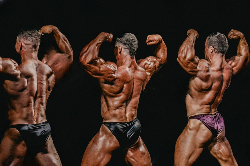 Three male bodybuilders in a back double biceps pose on stage showing deep, even competition tan